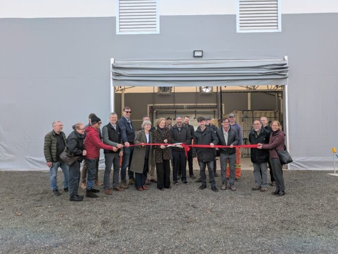 Group of people gather outside of facility holding ribbon, ready to cut it to mark opening